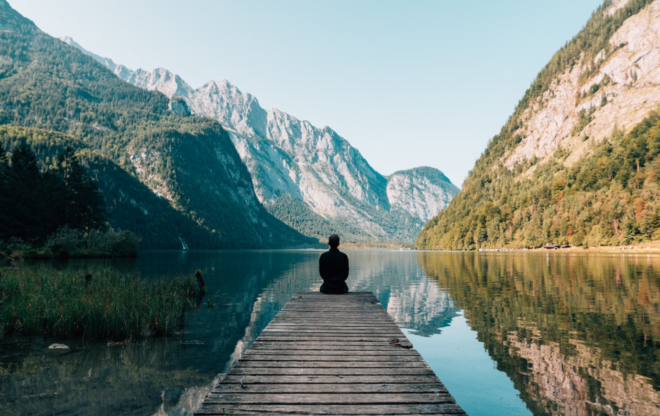 person sitting at the edge of a peer staring at water and mountains